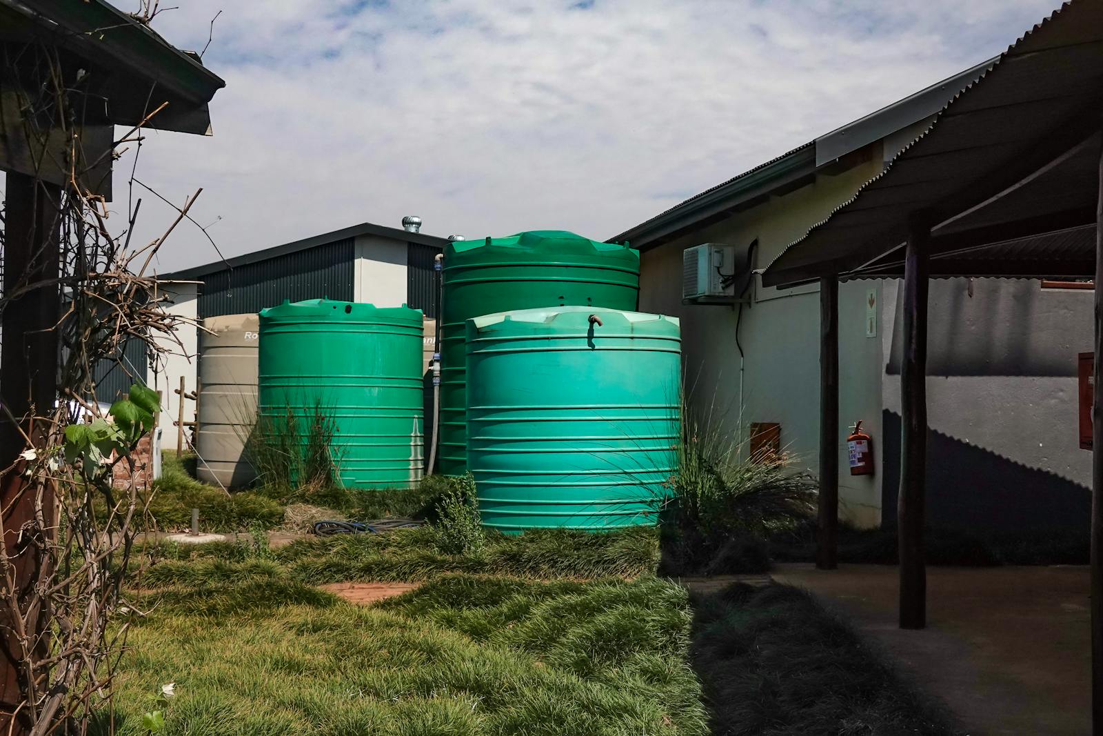 Three green water tanks beside a farmhouse in a rural setting under a cloudy sky.