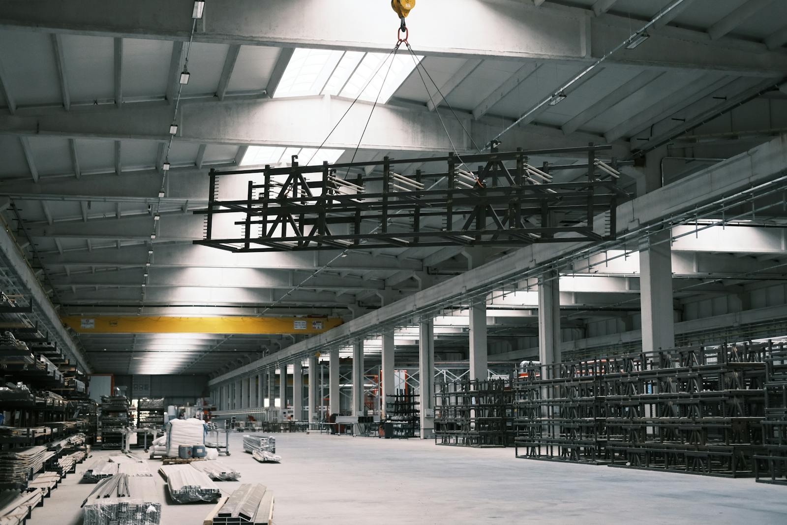 Spacious industrial warehouse interior with a yellow crane lifting metal framework.